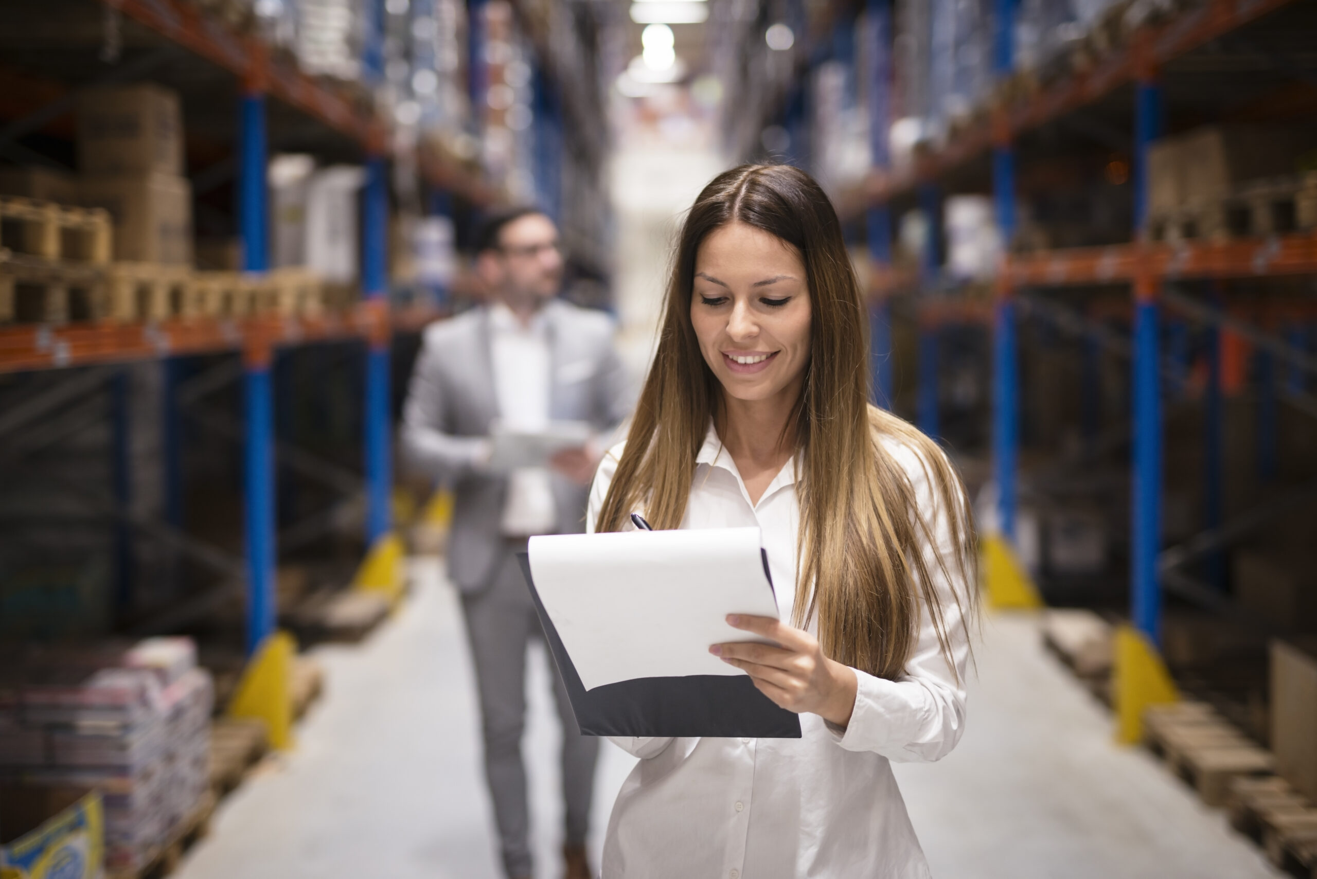 Portrait of gorgeous brunette woman manager controlling business in warehouse logistic center. Well dressed successful woman checking distribution while CEO walking behind her. Happy Business Owner in Warehouse